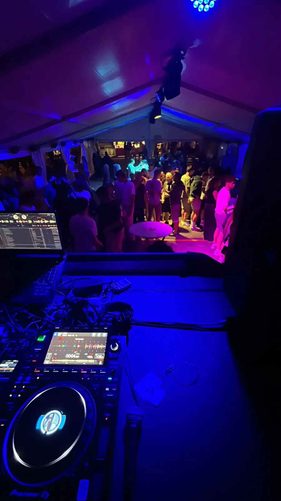 Vue depuis la régie DJ : contrôleur DJ au premier plan, tente éclairée en bleu et violet par les lyres et le public au loin, lors de l'édition 2025